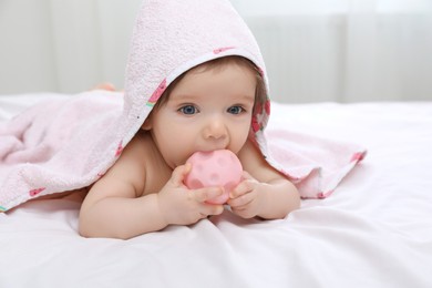 Cute little baby nibbling toy in hooded towel after bathing on bed at home