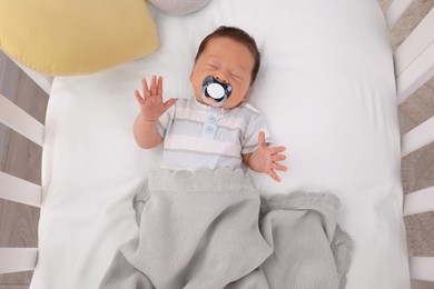 Photo of Cute newborn baby with pacifier sleeping under blanket in crib, top view. Bedtime