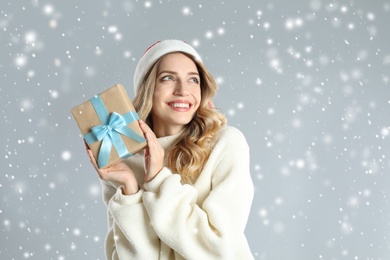 Beautiful young woman in Santa hat with Christmas present on light grey background