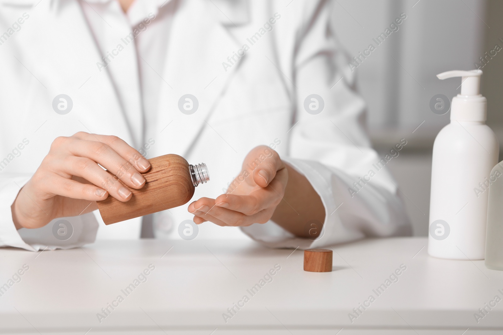 Photo of Dermatologist with bottle testing cosmetic product at white table indoors, selective focus