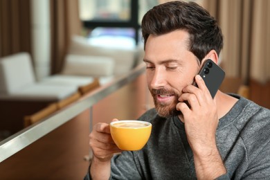 Photo of Handsome man with cup of coffee talking on phone indoors