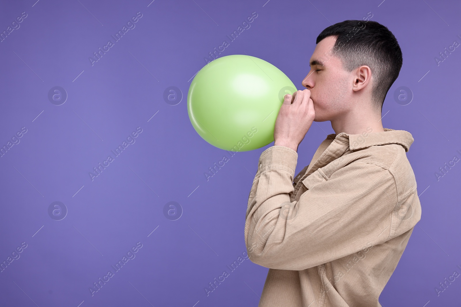 Photo of Young man inflating light green balloon on purple background. Space for text