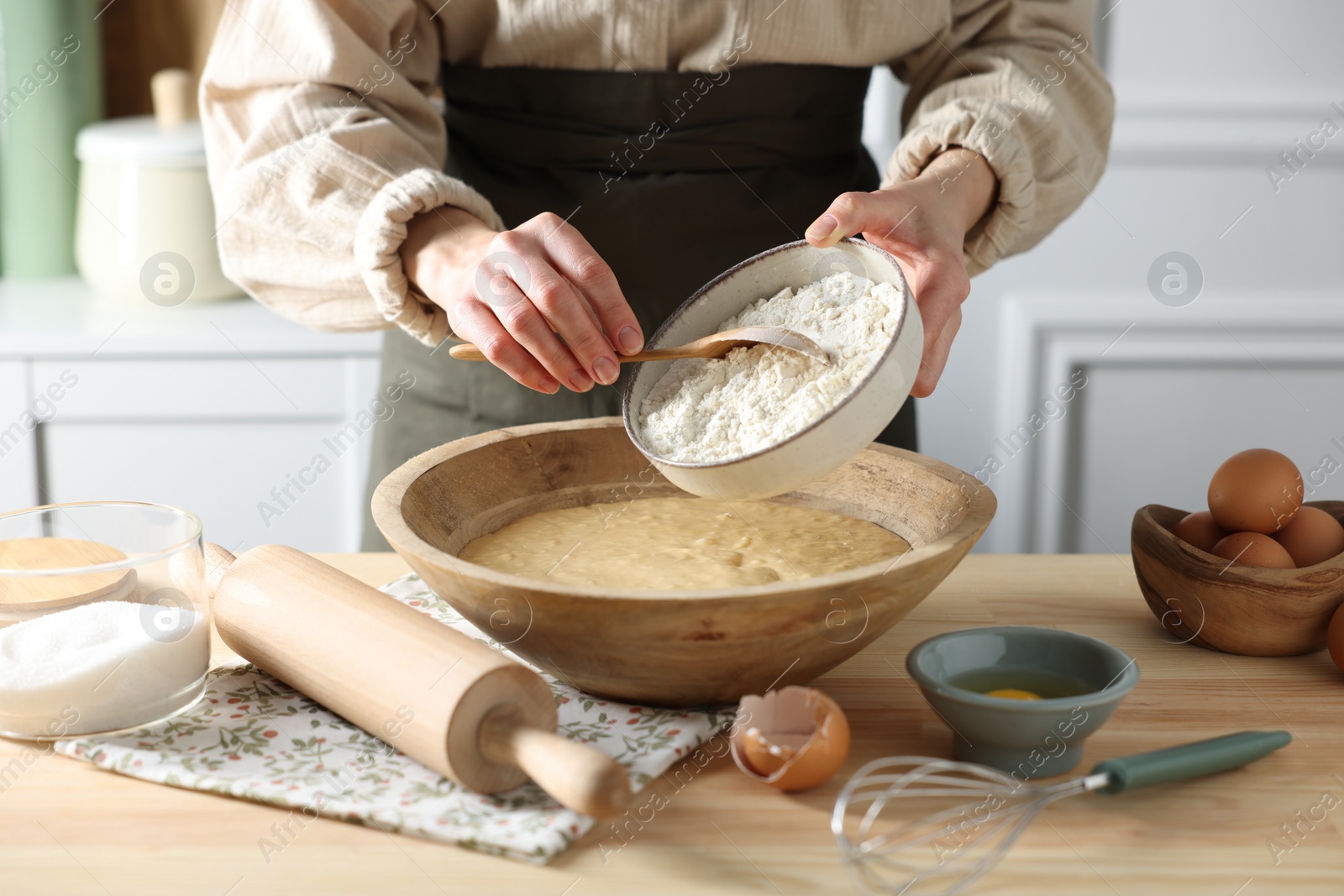 Photo of Making dough. Woman adding flour into bowl at wooden table in kitchen, closeup