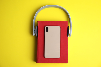 Book, modern headphones and smartphone on yellow background, top view