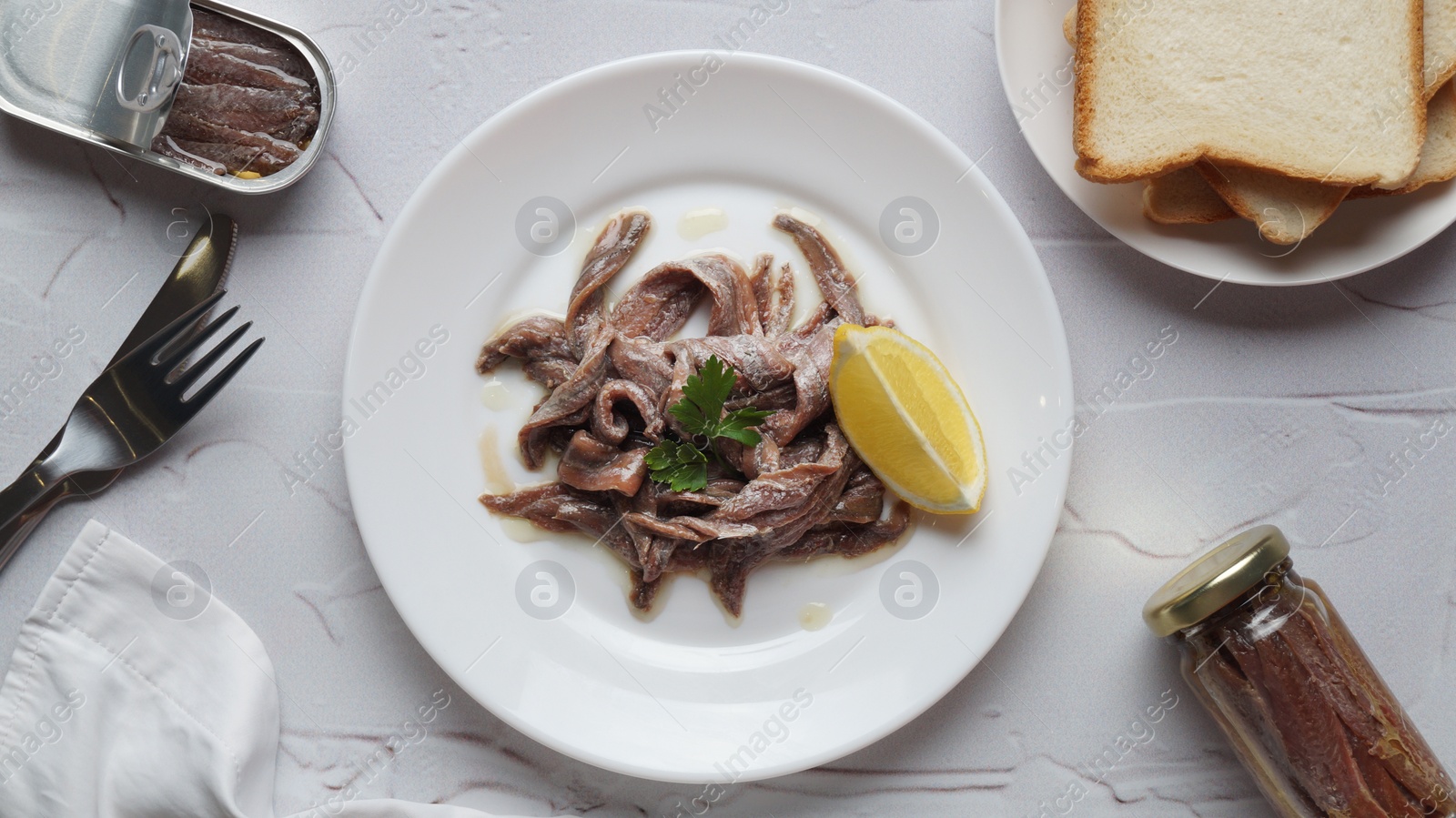 Photo of Plate with canned anchovy fillets, lemon wedge and pieces of toast on white textured table, flat lay