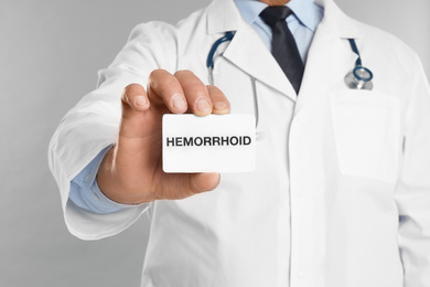 Photo of Doctor holding business card with word HEMORRHOID on light grey background, closeup