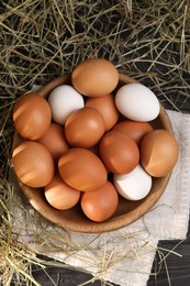 Photo of Fresh chicken eggs in bowl and dried hay on black wooden table, flat lay
