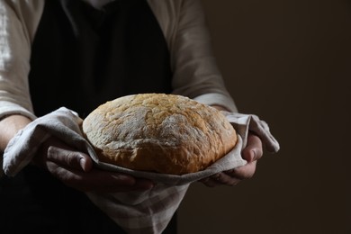 Photo of Man holding loaf of fresh bread on dark brown background, closeup