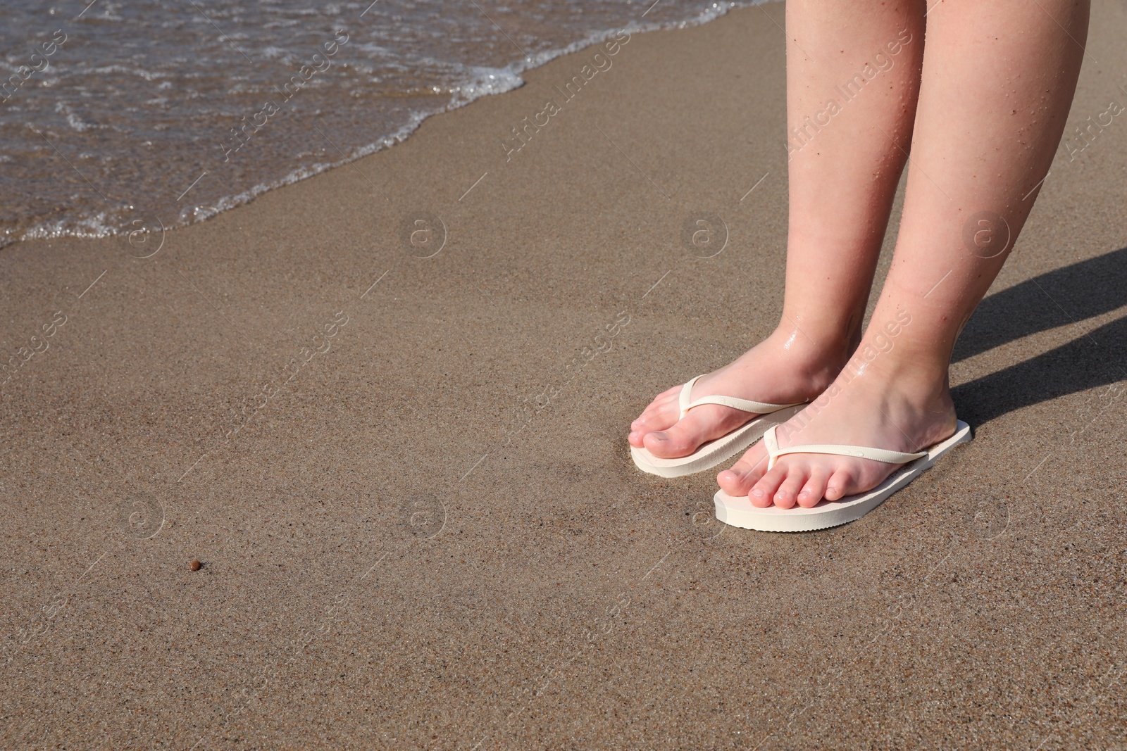 Photo of Woman in stylish white flip flops on sandy beach near sea, closeup. Space for text