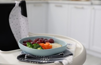 Photo of High chair with food in baby tableware on tray indoors, closeup