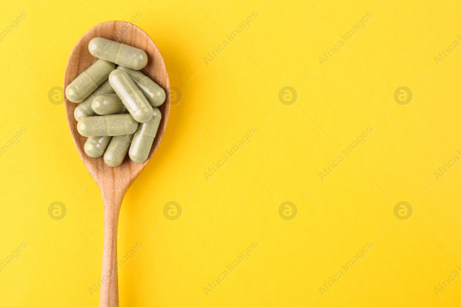Photo of Vitamin pills in wooden spoon on yellow background, top view. Space for text