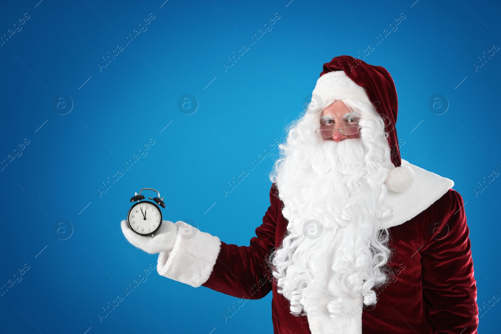 Photo of Santa Claus holding alarm clock on blue background, space for text. Christmas countdown