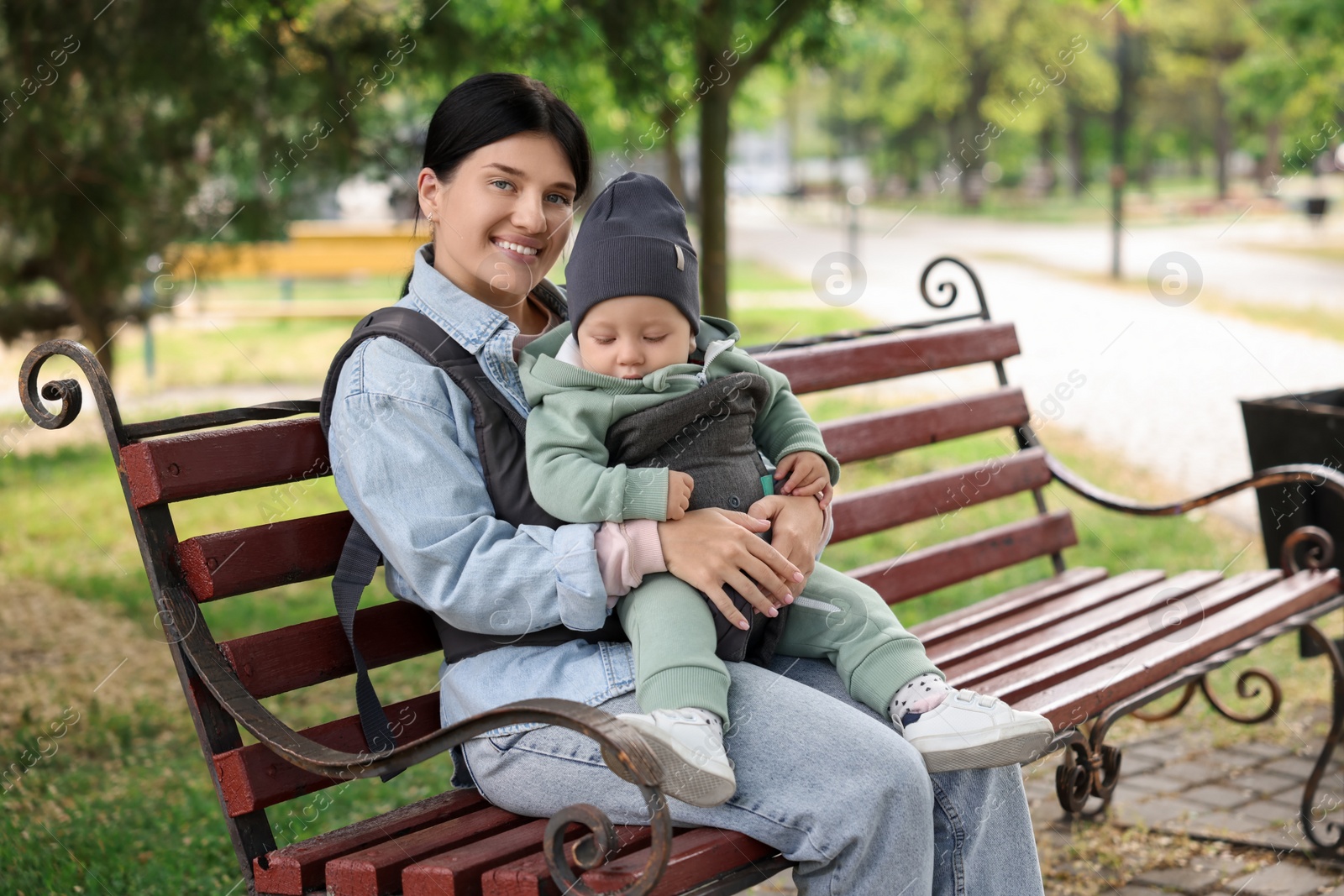 Photo of Mother holding her child in sling (baby carrier) on bench in park