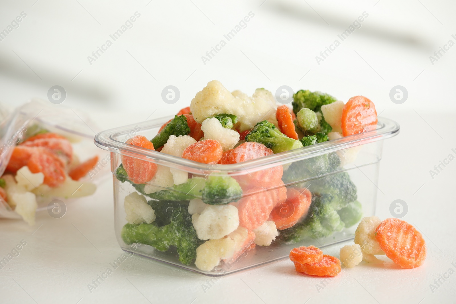 Photo of Mix of different frozen vegetables in plastic container on white textured table, closeup