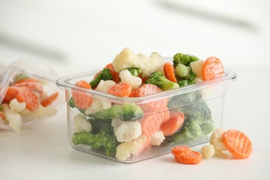 Mix of different frozen vegetables in plastic container on white textured table, closeup