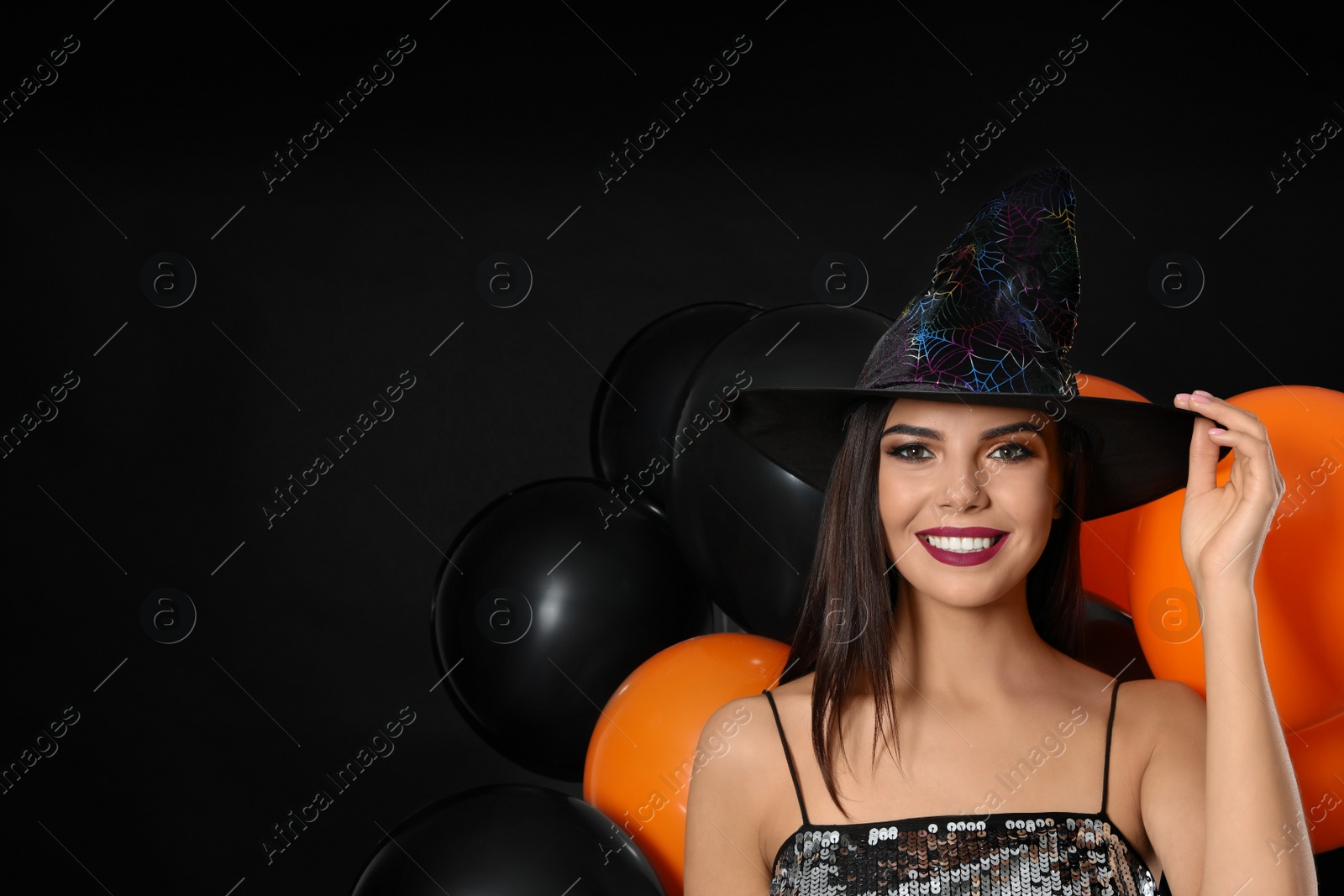 Photo of Beautiful woman wearing witch costume with balloons for Halloween party on black background, space for text