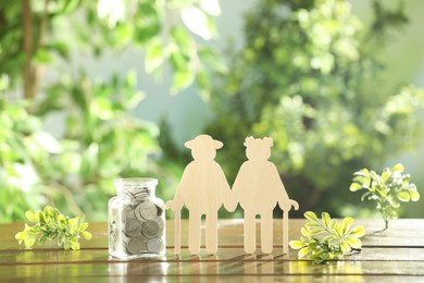 Photo of Pension savings. Figure of senior couple, coins in glass jar and green twigs on wooden table outdoors