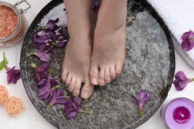 Photo of Woman soaking her feet in bowl with water and flowers on floor, top view. Spa treatment