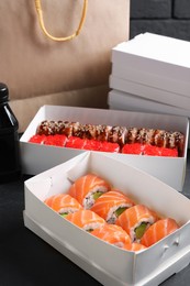 Photo of Food delivery. Boxes with delicious sushi rolls, paper package and soy sauce on black table