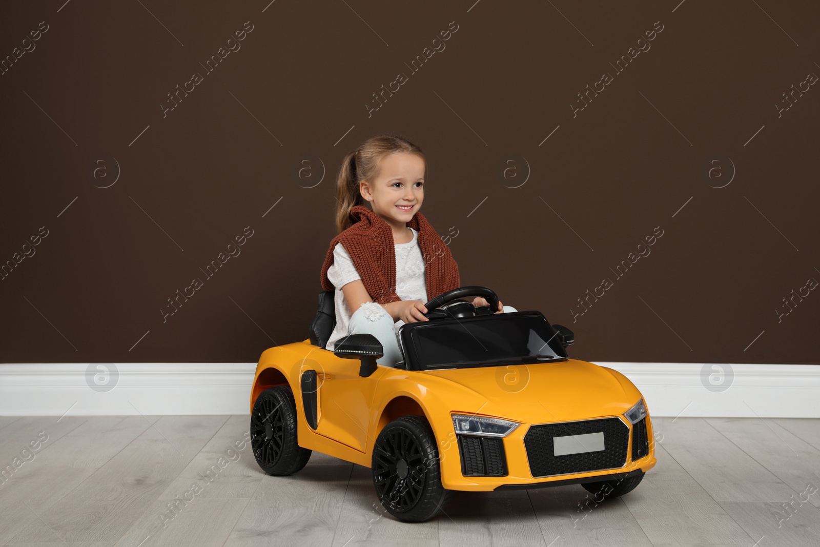 Photo of Cute little girl driving children's electric toy car near brown wall indoors