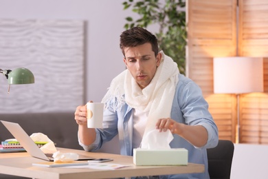 Photo of Sad exhausted man with tissue and cup of hot drink suffering from cold while working with laptop at table