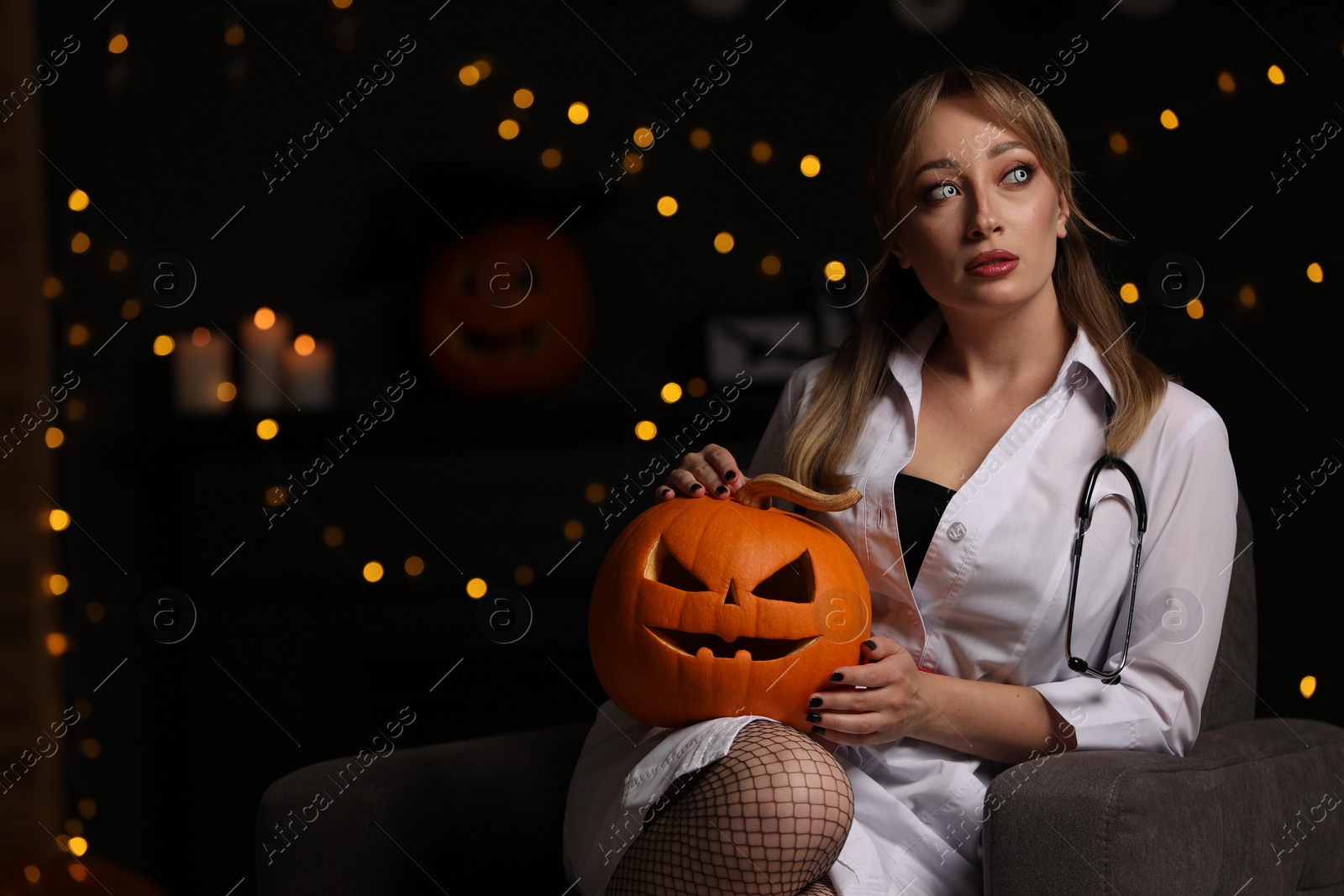 Photo of Woman in scary nurse costume with carved pumpkin against blurred lights indoors, space for text. Halloween celebration