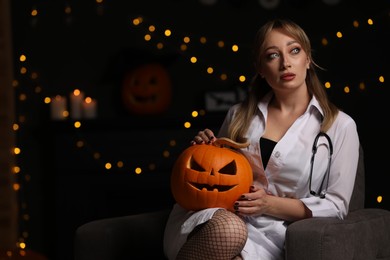 Photo of Woman in scary nurse costume with carved pumpkin against blurred lights indoors, space for text. Halloween celebration