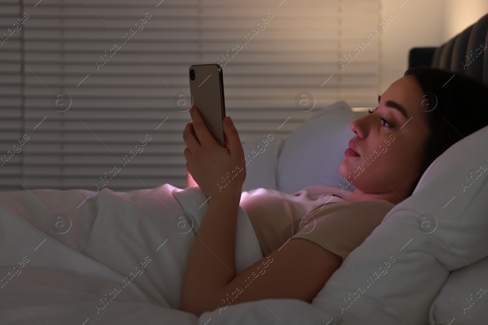 Photo of Young woman using modern smartphone in bed at night. Internet addiction