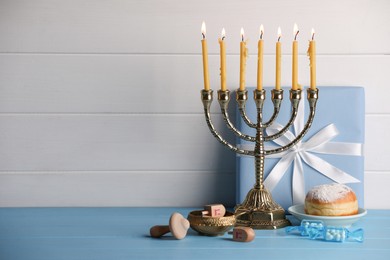 Photo of Hanukkah celebration. Menorah with burning candles, dreidels, donut and gift box on light blue wooden table, space for text