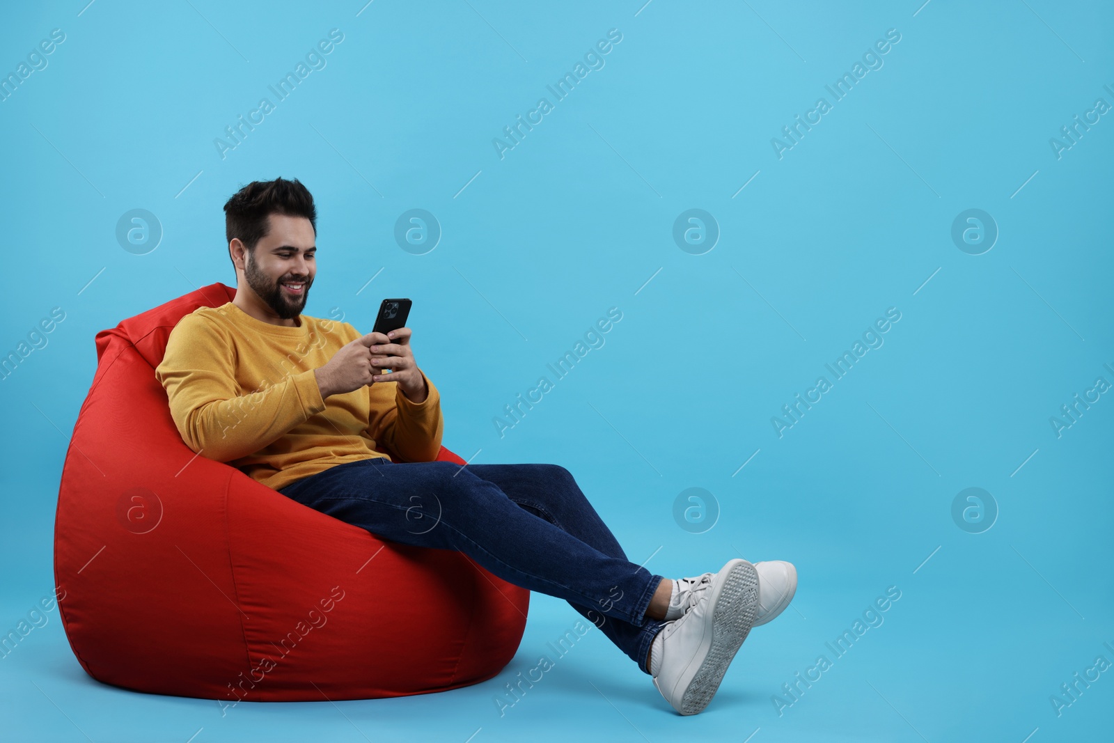 Photo of Happy young man using smartphone on bean bag chair against light blue background. Space for text