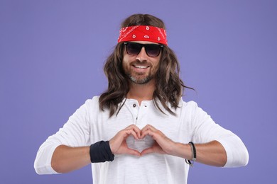 Photo of Stylish hippie man in sunglasses making heart with hands on violet background