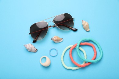 Stylish sunglasses, bracelets, seashells and rings on light blue background, flat lay