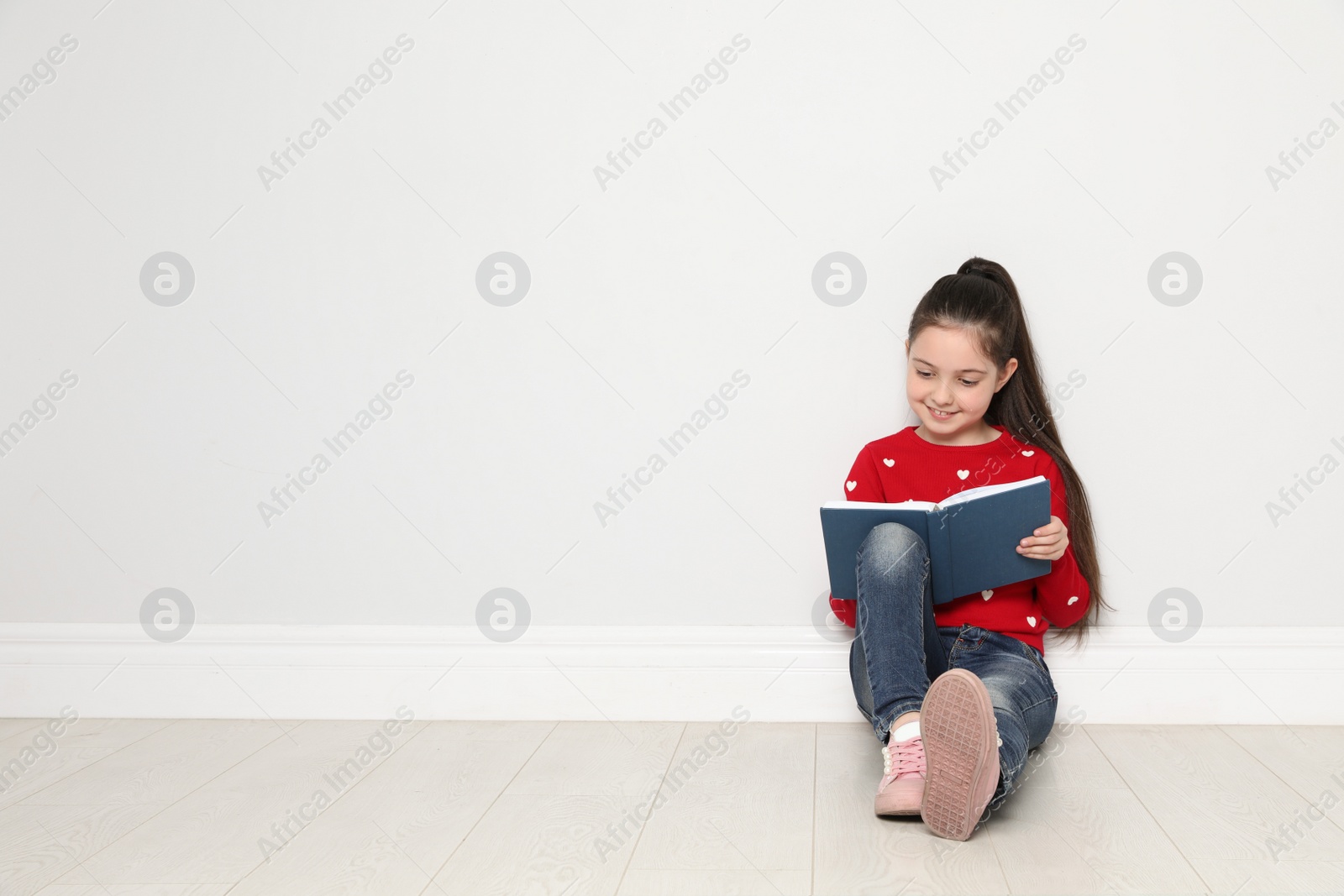 Photo of Cute little girl reading book on floor near white wall, space for text