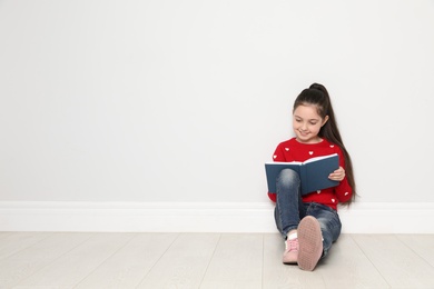 Cute little girl reading book on floor near white wall, space for text