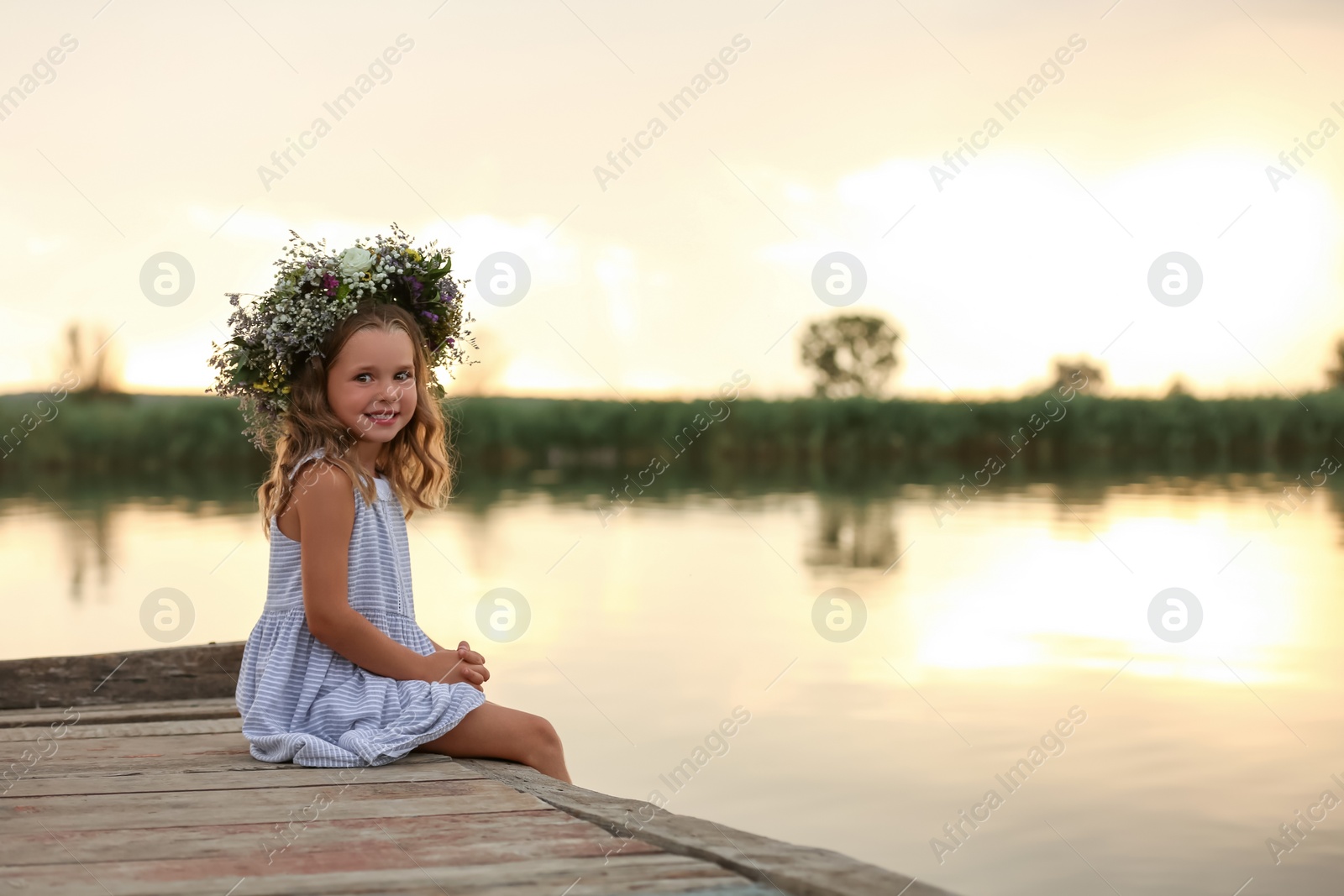 Photo of Cute little girl wearing wreath made of beautiful flowers on pier near river