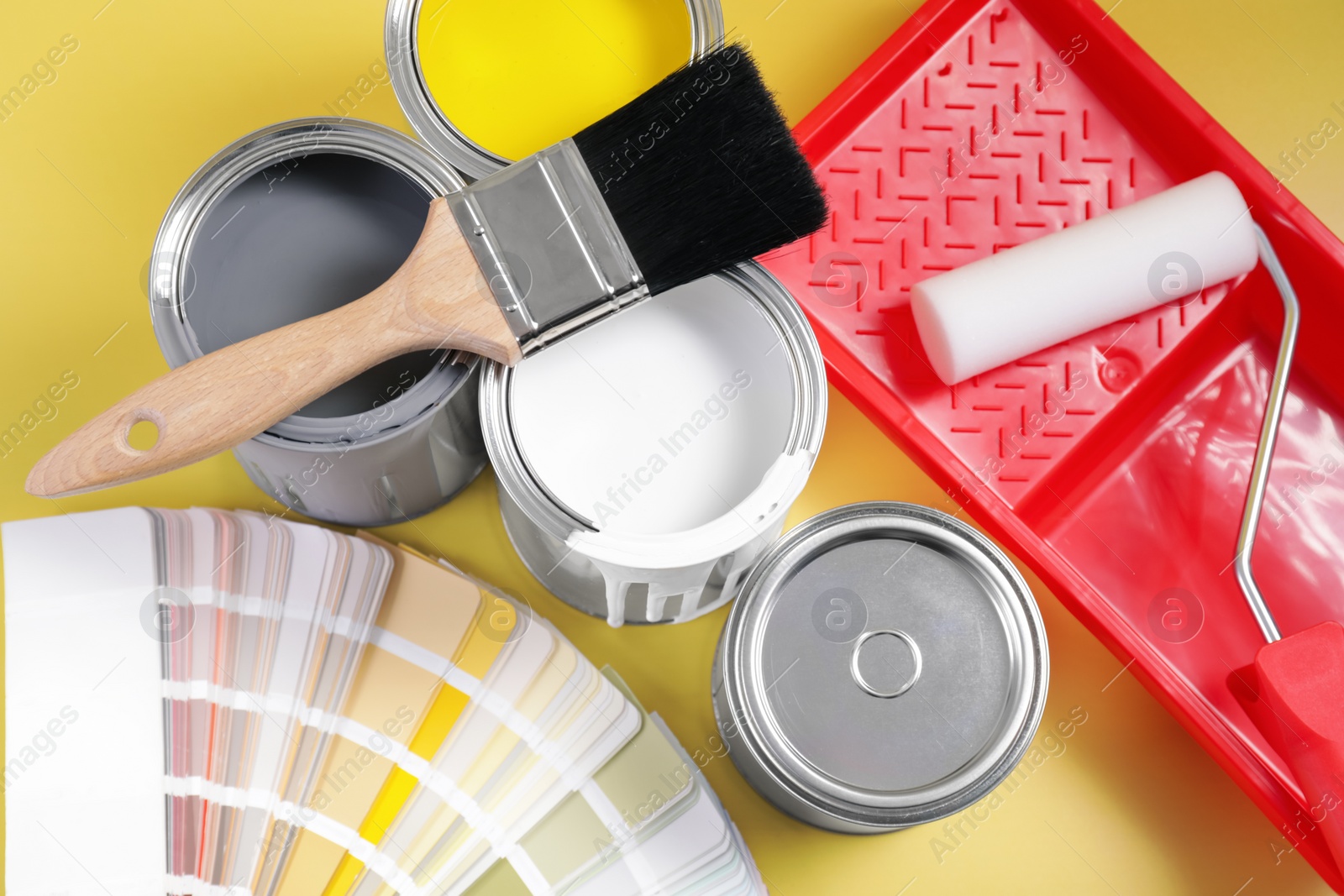 Photo of Cans of paints, brush, roller, tray and palette on yellow background, flat lay