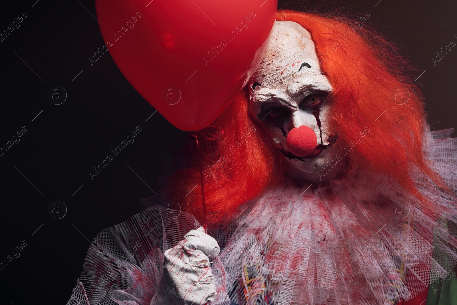 Photo of Terrifying clown with red air balloon on black background. Halloween party costume