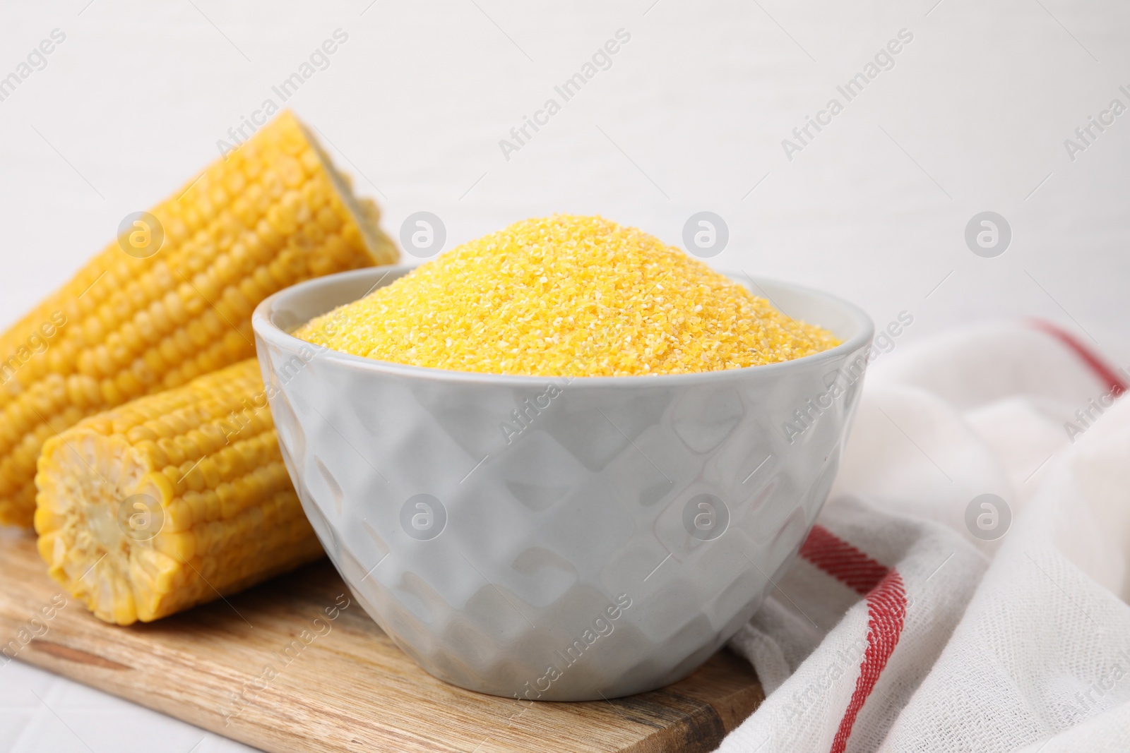 Photo of Raw cornmeal in bowl and corn cobs on white table, closeup