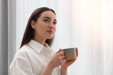 Photo of Beautiful woman with cup of drink near window at home, space for text. Lazy morning