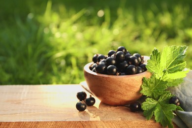 Ripe blackcurrants in bowl and leaves on wooden surface outdoors. Space for text