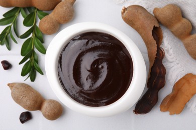 Photo of Bowl of tasty tamarind sauce, leaves and fresh pods on white table, flat lay