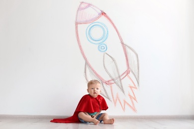 Adorable little child playing astronaut near wall with drawing of spaceship indoors