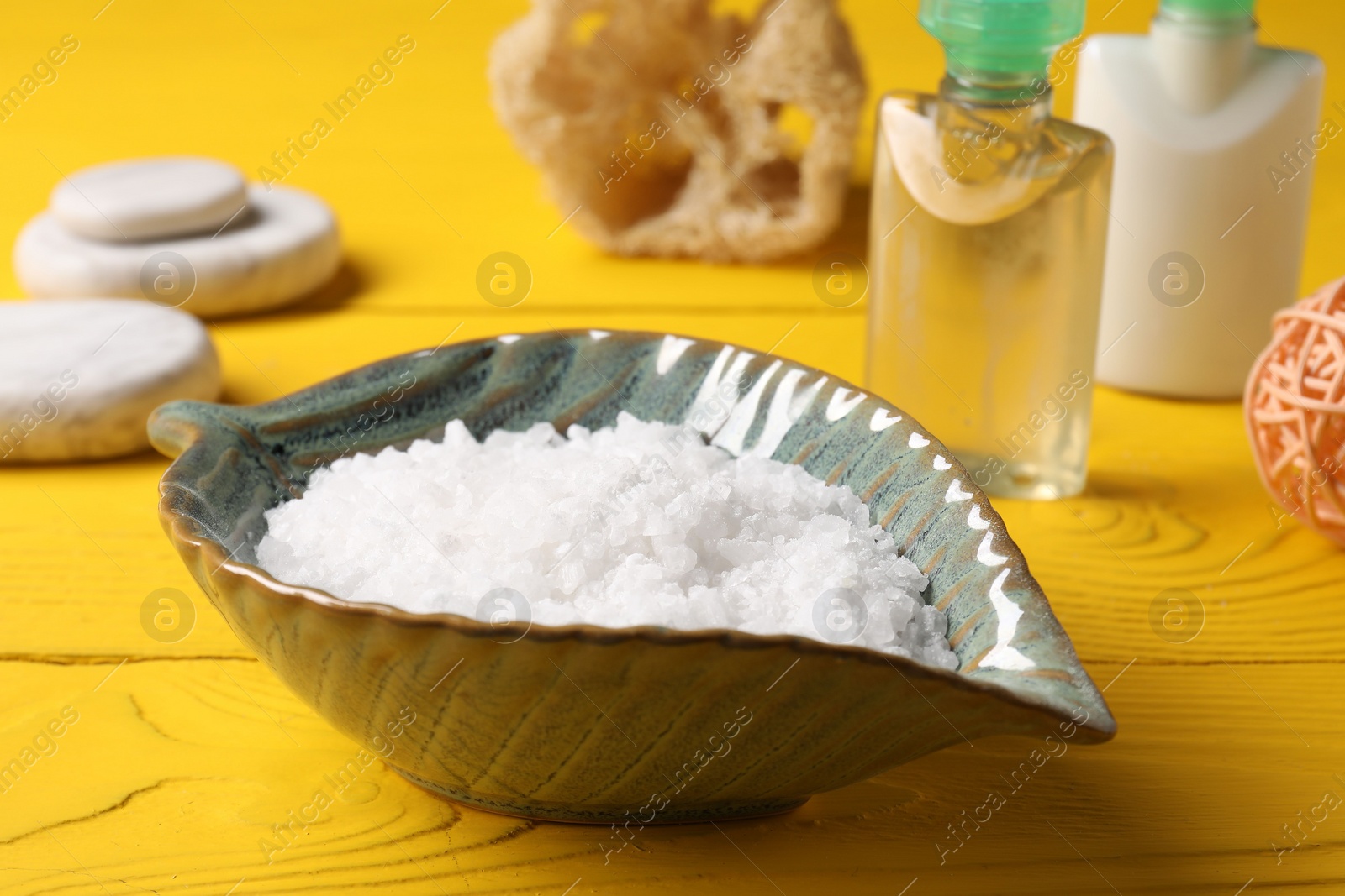 Photo of Natural sea salt in bowl and other spa products on yellow wooden table, closeup