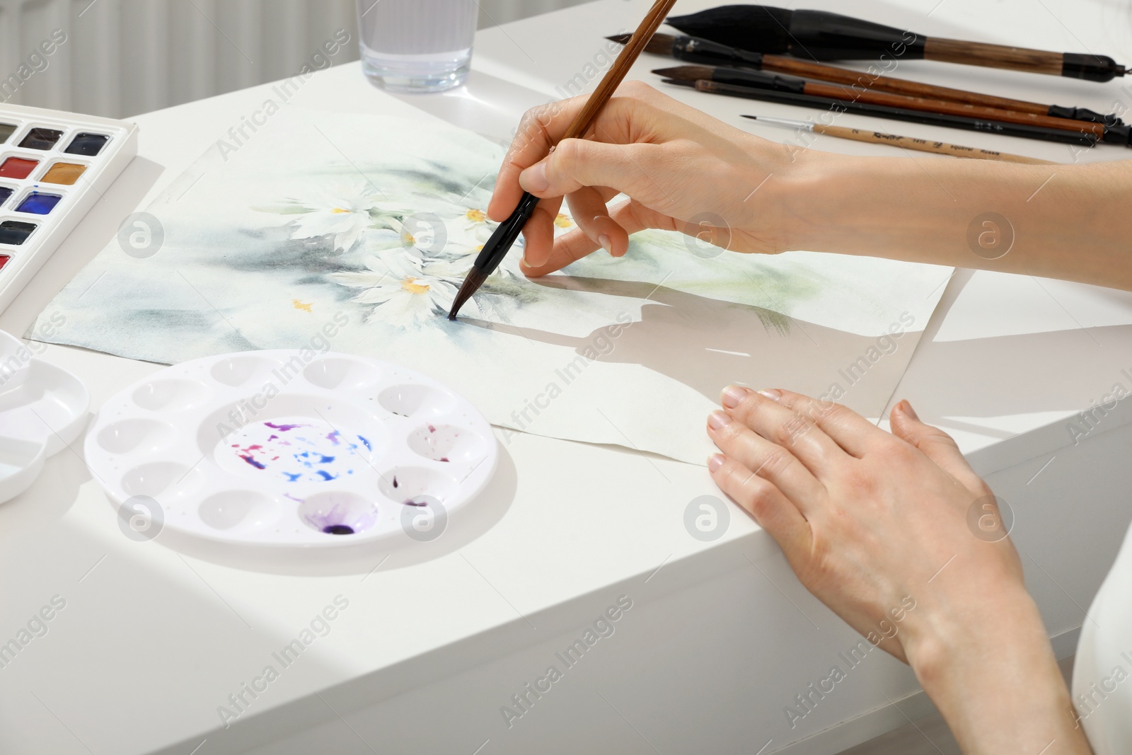 Photo of Woman painting flowers with watercolor at white table indoors, closeup. Creative artwork