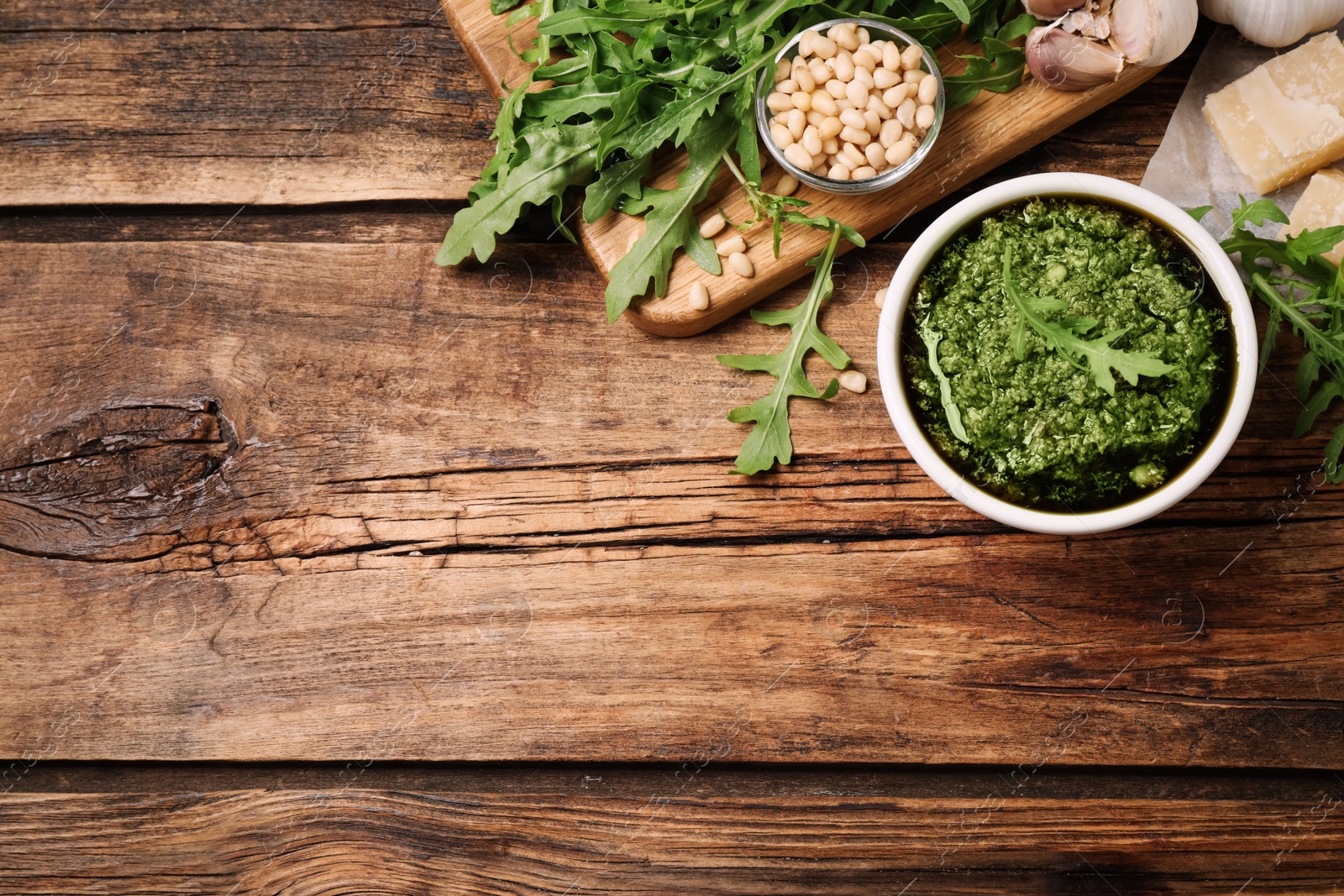 Photo of Bowl of tasty arugula pesto and ingredients on wooden table, flat lay. Space for text