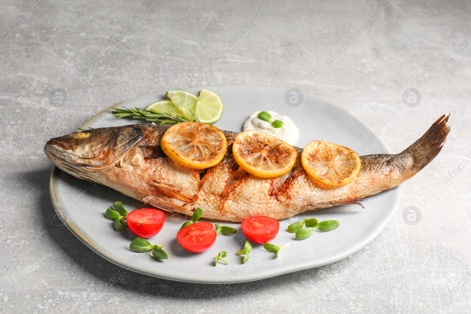 Photo of Delicious roasted sea bass fish served with lemon, tomatoes and sauce on light grey table