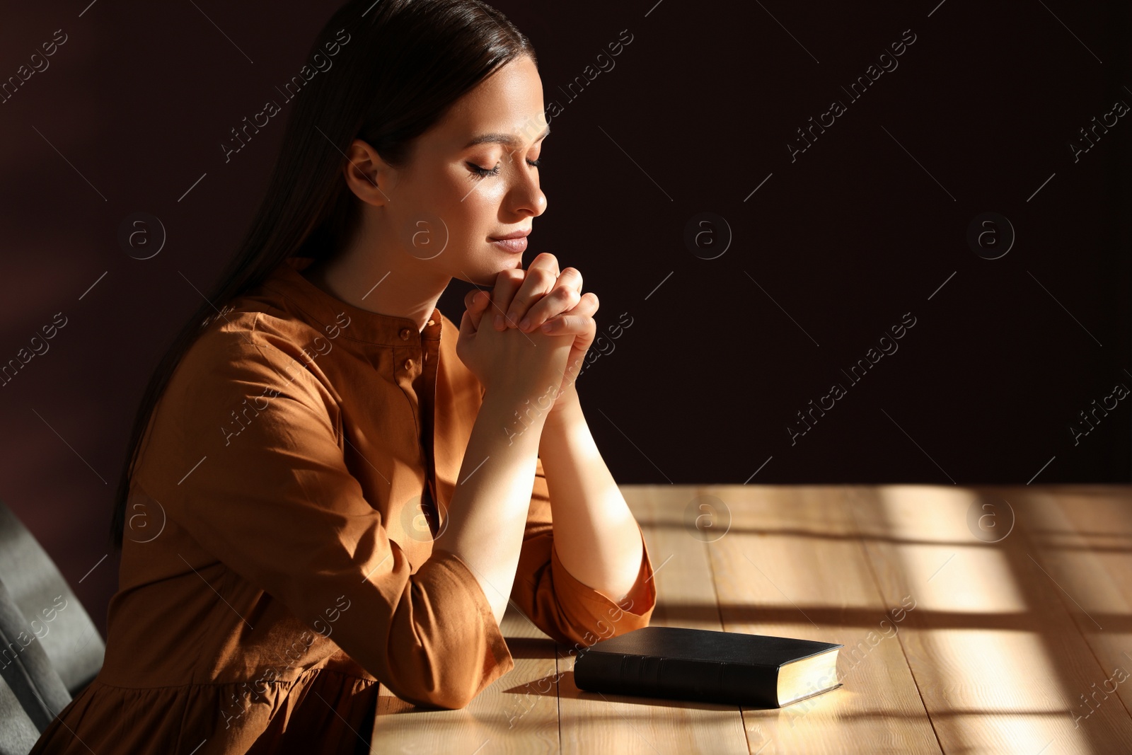 Photo of Religious young woman praying over Bible at table indoors