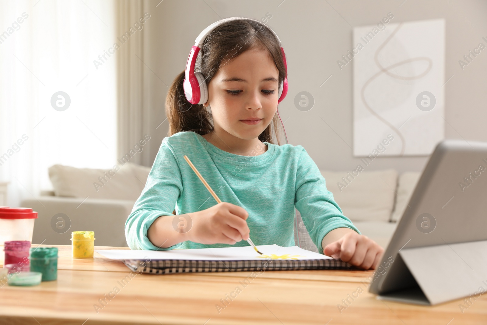 Photo of Little girl with headphones drawing on paper at online lesson indoors. Distance learning