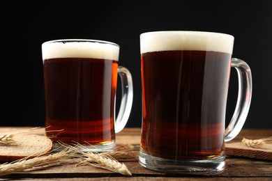 Photo of Mugs of delicious kvass, spikes and bread on wooden table against black background