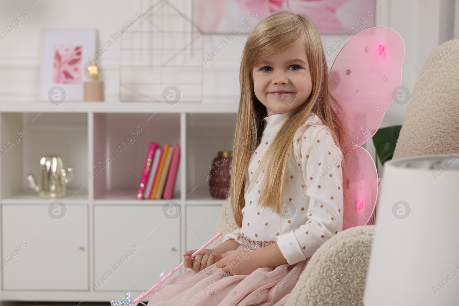 Photo of Cute little girl in fairy costume with pink wings and magic wand in armchair at home, space for text